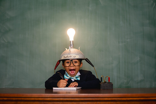 A young nerd boy has a bright idea with copy space in front of a chalkboard. His lightbulb helmet helps him think of the next big idea.