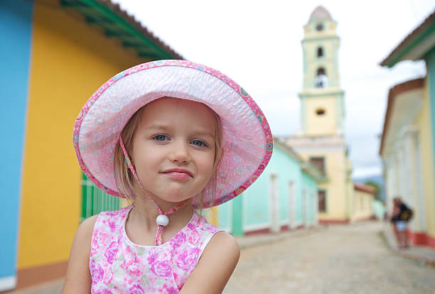 A young girl poses with her hands cupped under her chin, smiling at the camera. She wears a blue tank top and stands in front of a weathered blue wooden wall or shutters. Red string lights hang in the background, slightly out of focus. The portrait uses selective focus to highlight the child's expression while creating a rustic, atmospheric setting.