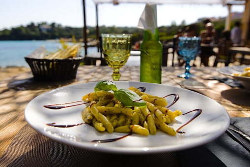 Penne pasta on a white plate in Sardinia, with pesto, balsamic vinegar, and fresh basil leaves.