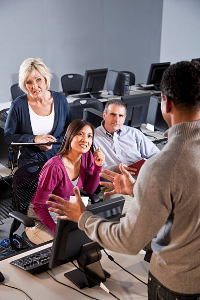 A rear view of adults is shown during a computer class.  The man teacher is in the background, blurred and pointing to the white wall behind him.  The students are sitting at large computer monitors, with either white or blue-gray screens.  There are both woman and men in the room.