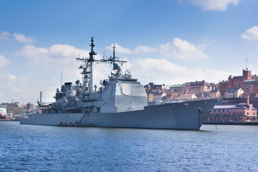 A modern warship is anchored in port with a European city in the background (Stockholm)