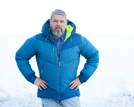Man, 56 years old with gray hair and a gray beard, stands in front of a snowy area in winter, facing the camera, hands on hips, critical look, thick down jacket, down jacket, blue shirt