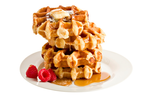 A stack of four Belgian waffles and three luscious red raspberries on a white plate with a melting slab of butter and maple syrup still barely trickling onto the butter. Isolated on white
