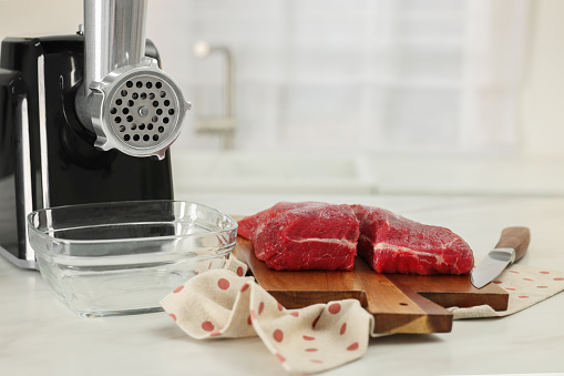 Electric meat grinder with beef and knife on white marble table in kitchen