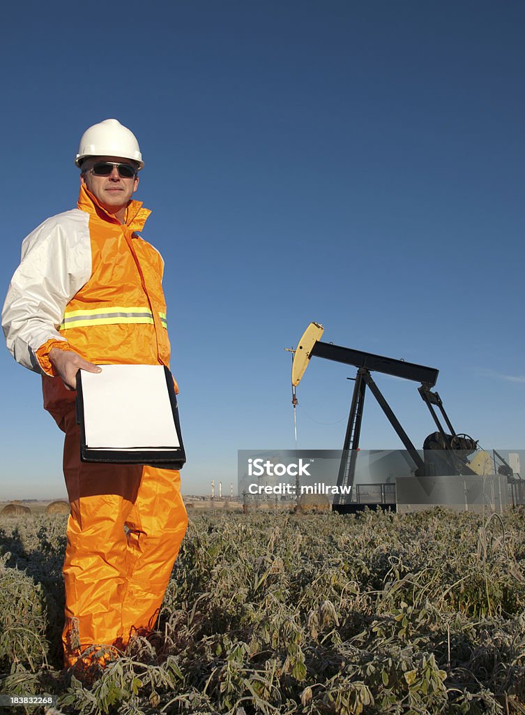 Oil Worker in Safety Gear at Well Pumpjack Soft early morning sun. Alberta Canada in the fall.Suitable for a variety of themes: - balance of industry and environment; - Linkage between oil and gas and the petrochemical business (plant in backgroun)- the need for regulation and monitoring (authority figure with blank clipboard) Agriculture Stock Photo
