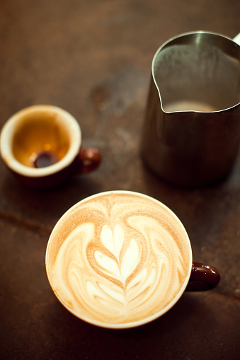 A freshly poured hot espresso drink on a counter top, the milk / cream foam art poured in the shape of a heart. Visible behind are a steel pitcher of steamed milk foam and the mug that the espresso shot was pulled in.  Vertical with copy space.