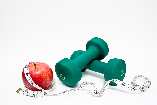 Red apple with measuring tape with green hand weights on white background indicating the importance of both healthy eating and fitness to stay in shape and for overall health and wellbeing