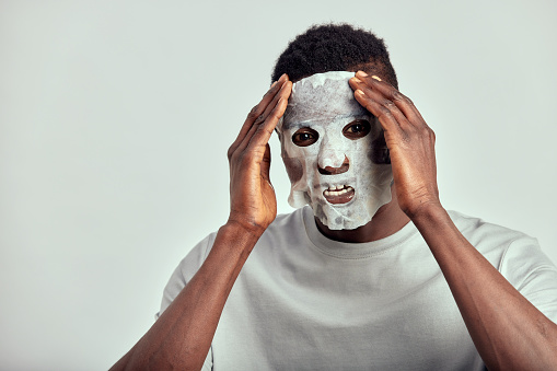 Studio portrait of young smiling black man applying moisturizing paper mask on his face. Happy African American millennial man uses beauty products in his skincare routine to maintain healthy look.