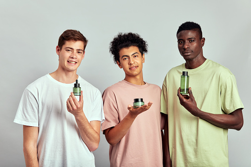 Three young handsome men of different races with cosmetic products in their hands. Caucasian, Hispanic and African American millennials practice skincare routine to keep their young and healthy look.
