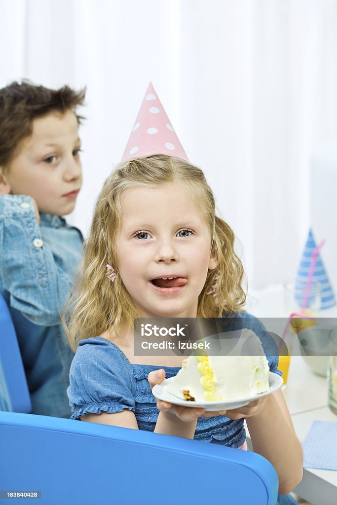 Cute girl with cake Kids at a birthday party. Focus on the cute little girl wearing party hat and blue dress showing a tasty piece of birthday cake to camera and licking her lips. 6-7 Years Stock Photo