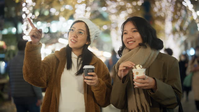 Two Asian women are walking and having fun talking in the city at night with many Christmas lights in the background.