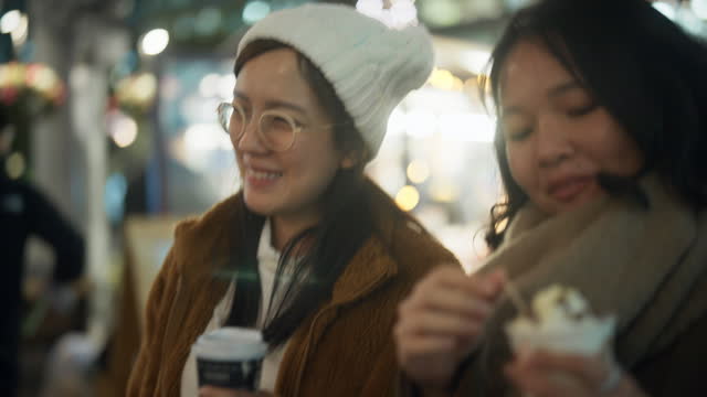 Two Asian women are walking and having fun talking in the city at night with many Christmas lights in the background.