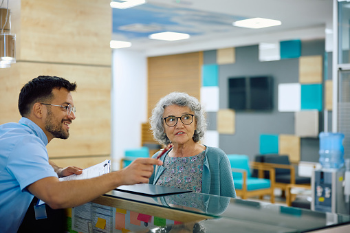 Happy medical technician talking to female senior patient at reception desk at doctor's office. Copy space.