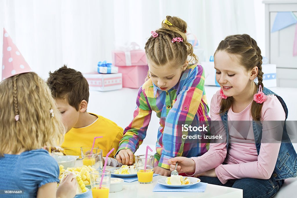 Kids birthday party Birthday party. Four cute kids sitting at the table and eating a birthday cake. Many gift boxes in the background. 6-7 Years Stock Photo