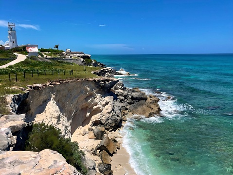 A rocky shoreline in Cancun, Mexico. The water is light blue and there are large waves. There is a white lighthouse and an old shack in the background. There is a cliff with grass growing and a fence at the top. The sky is blue with small clouds.