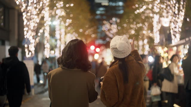 Rear view of two Asian women having fun walking and talking in the city at night with many Christmas lights.