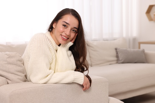 Beautiful young woman in stylish warm sweater on sofa at home, space for text