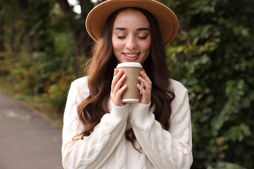 Beautiful young woman in stylish warm sweater holding paper cup of coffee outdoors