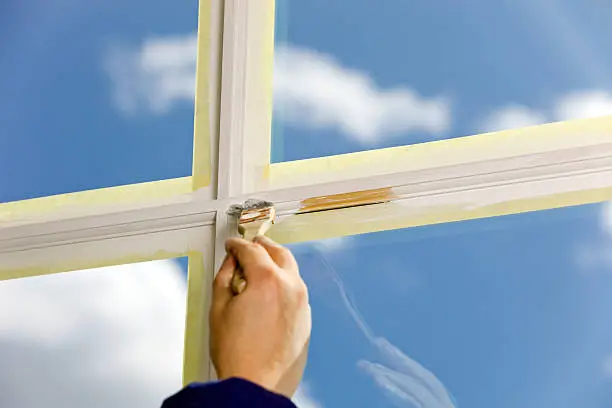 A housepainter is using a small paint brush to paint a cross section of window trim with blue sky and clouds on the other side of the glass. A fluorescent light is slightly reflected in the upper right of the glass. Focus is on the left of the unpainted area. The brush and hand are blurred due to motion and depth of field.