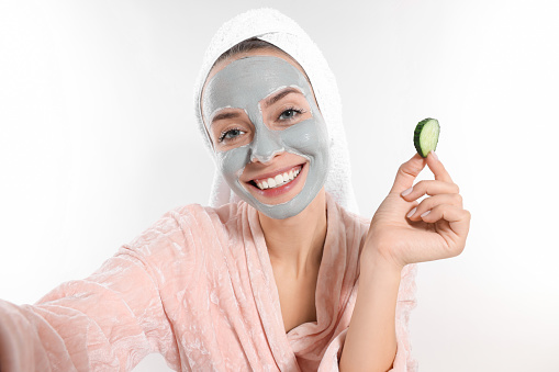 Woman with face mask and cucumber slice taking selfie on white background. Spa treatments