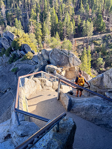 Adult white man, with backpack on his shoulders, standing on the narrow steps ascending to the Moro Rock tourist viewpoint in Sequoia National Park. With a gaze, he absorbs the breathtaking sight of the towering trees that surround him