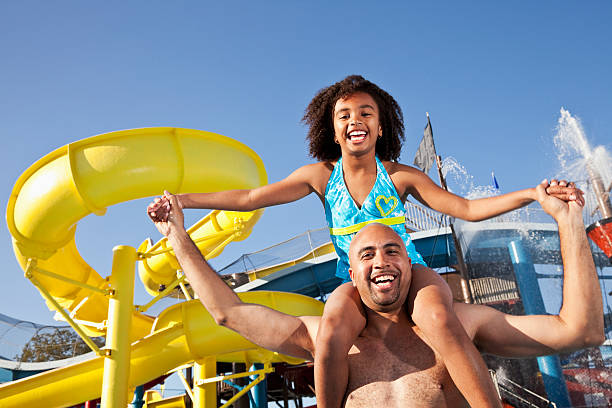 Father and daughter in front of yellow water slide A young girl wearing a blue bikini smiles as she sits on her father's shoulders at an amusement park.  The shirtless father smiles while holding her arms with a yellow water slide in the background. amusement park photos stock pictures, royalty-free photos & images