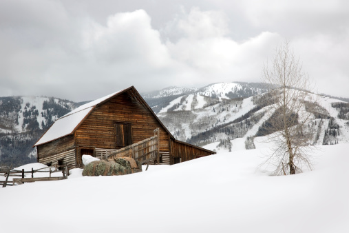 Aged barn on snowy hillside with ski lifts and ski slopes in background.
