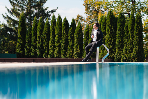 Handsome businessman in suit and with sunglasses sitting on the edge of a swimming pool and looking away at suny summer day