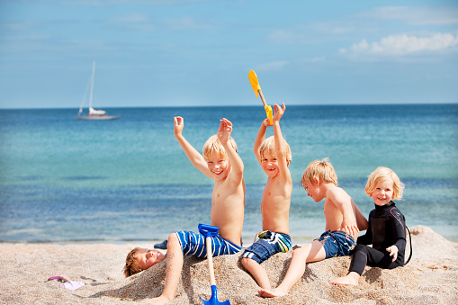 Group of happy young children sitting on another boy who they have buried in the sand. At the beach with the sea in the background in summer. Taken in Cornwall, UK. 