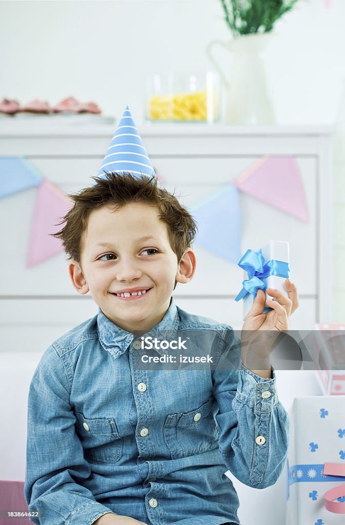 Cute boy with birthday present "Kids Birthday Party. Cute little boy wearing party hat holding small gift box in his hand, looking at it and smiling." 6-7 Years Stock Photo