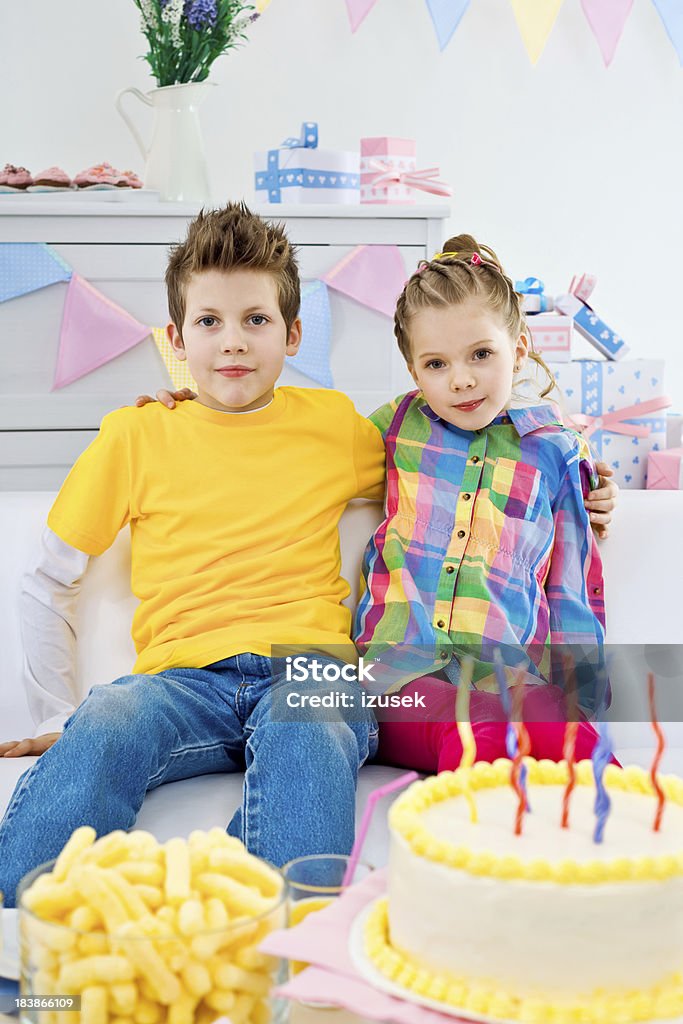 Siblings at a birthday party "Brother and sister having birthday party, sitting on sofa in domestic room, looking at camera and smiling. Birthday cake in the foreground, lots of gifts in the background." 6-7 Years Stock Photo
