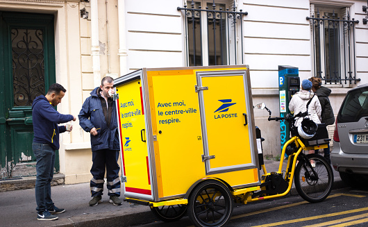 Paris, France: Two postal workers chat next to a postal delivery tricycle. The delivery tricycle has a message: Avec moi, le centre-ville respire (With me, downtown can breathe.)