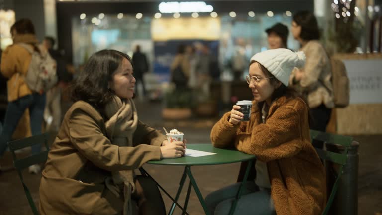 Asian friends chatting and drinking hot drinks at a sidewalk cafe table in the city at night. During the Christmas holidays