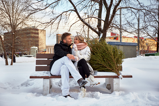 Beautiful young couple in love sitting on a bench at Christmas market and just bought a Christmas tree. Man and woman are preparing for Christmas. Anticipation of the New Year Eve in a snowy city.