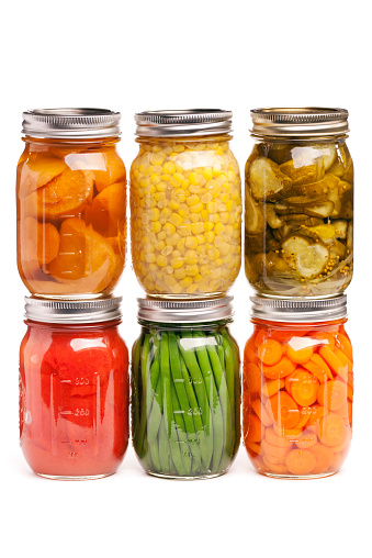 A variety of canned vegetables lined up in rows of glass jar containers. Food staples canned as homemade preserves include slices of carrots, green beans, tomatoes, corn, sweet potatoes, and cucumber pickles. Isolated on a white background.