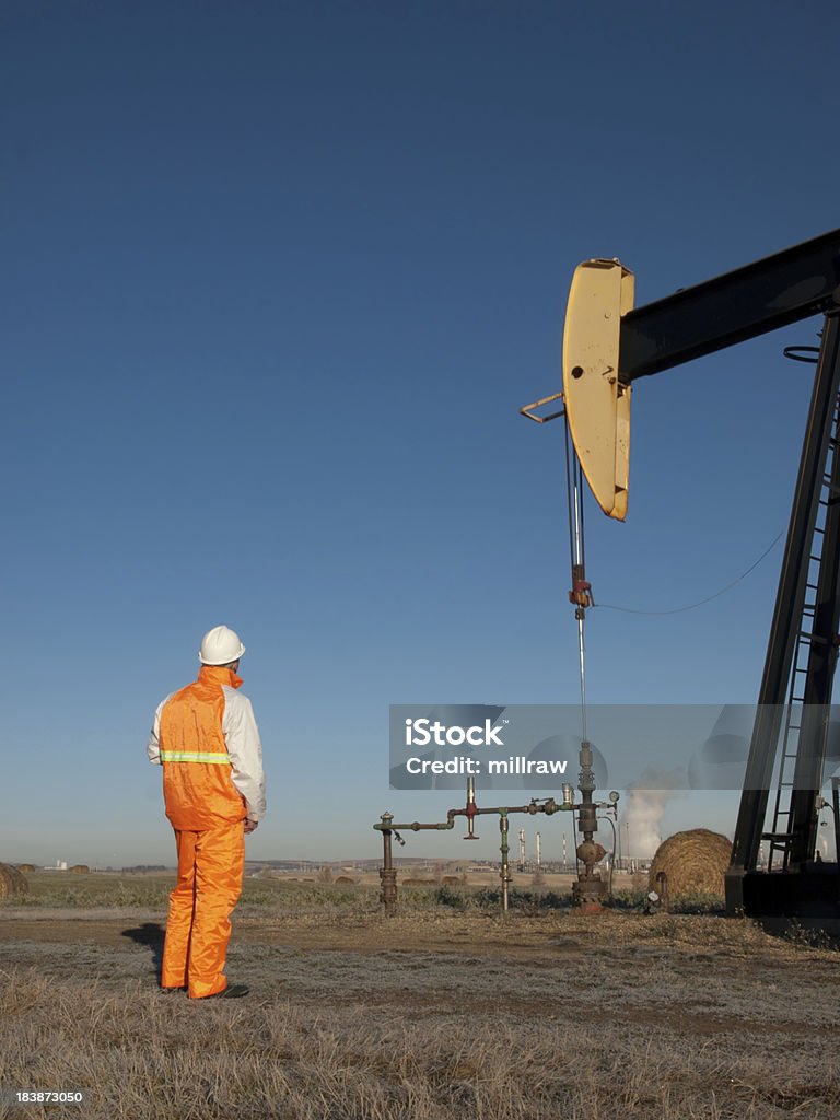 Oil Worker in Safety Gear at Well Pumpjack Soft early morning sun. Alberta Canada in the fall.Suitable for a variety of themes: - balance of industry and environment; - Linkage between oil and gas and the petrochemical business (plant in backgroun)- the need for regulation and monitoring (authority figure with blank clipboard) Adult Stock Photo