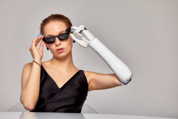 istock Young charming girl with disability wearing sensory bionic prosthetic arm sits at the table in studio. Beautiful woman in a black dress adjusting sunglasses freely controlling artificial robotic hand. 1838738263