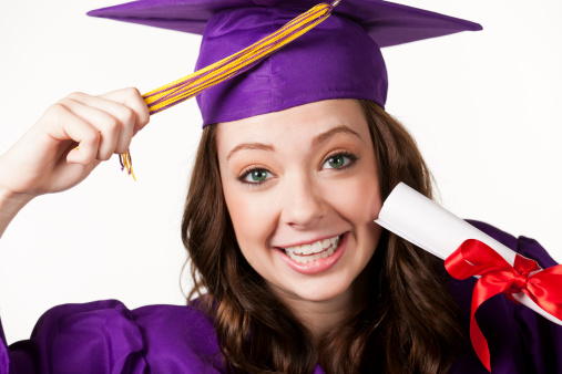 Portrait of a young woman graduate in a purple cap and gown holding her diploma and pulling her tassel from one side to the other while smiling happy at the camera.