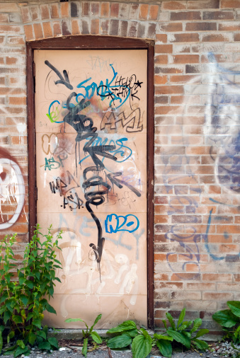 A graffiti filled back door on  a brick wall and with weeds growing out of the pavement.