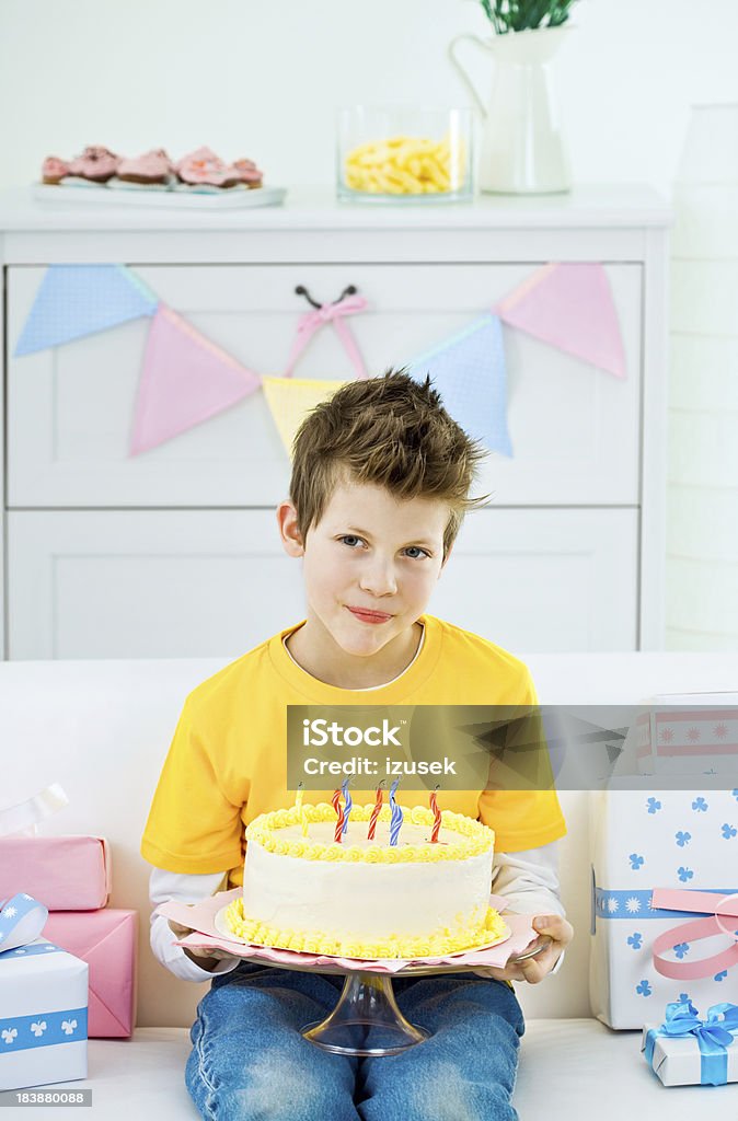 Cute boy with birthday cake Cute little boy wearing yellow t-shirt and jeans sitting on sofa among birthday presents and holding a birthday cake with candles in his hands. Looking away and smiling. 8-9 Years Stock Photo