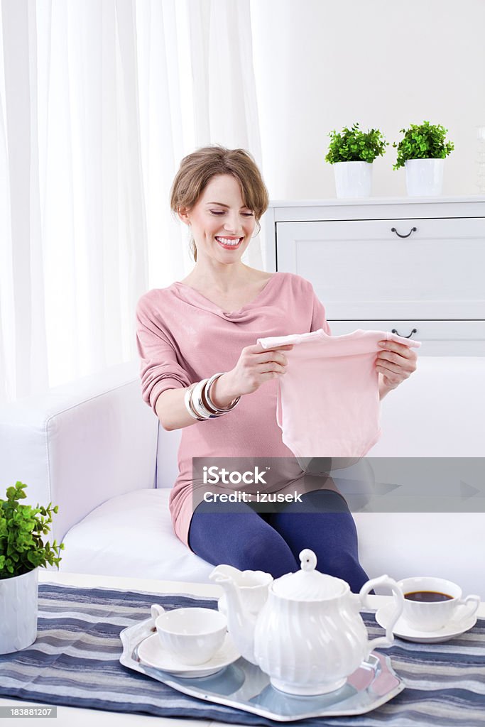 Happy pregnant woman "A beautiful, smiling pregnant young adult woman sitting on sofa in living room and looking at baby clothes." Only Women Stock Photo
