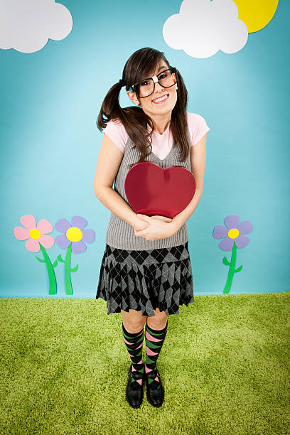 Happy, Nerdy Woman Holding Heart-Shaped Tin "Color photo of a nerdy young woman holding a tin heart while standing with a cheesy smile in a whimsical, outdoor world." embarrassment skirt photos stock pictures, royalty-free photos & images