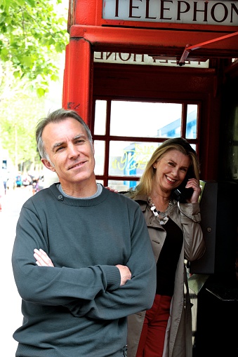 Man aged 50 to 60 leaning against red London telephone booth, smiling, arms crossed, woman aged 50 to 60 behind him speaking on the telephone