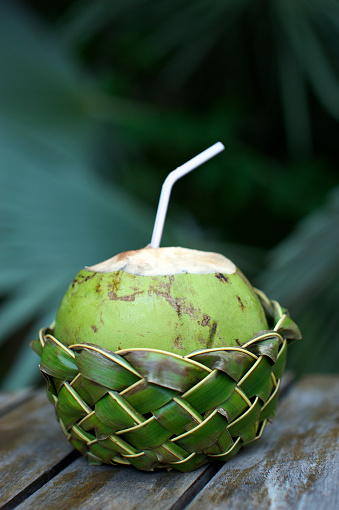 Fresh Brazilian coco gelado drinking coconut in woven palm basket rests on weathered wooden table against green jungle background