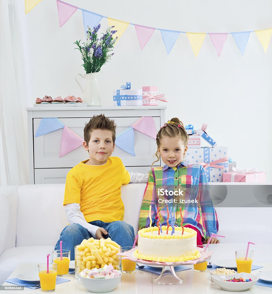Siblings at a birthday party "Brother and sister having birthday party, sitting on sofa in domestic room, looking at camera and smiling. Birthday table with a birthday cake in the foreground, lots of gifts in the background." 6-7 Years Stock Photo
