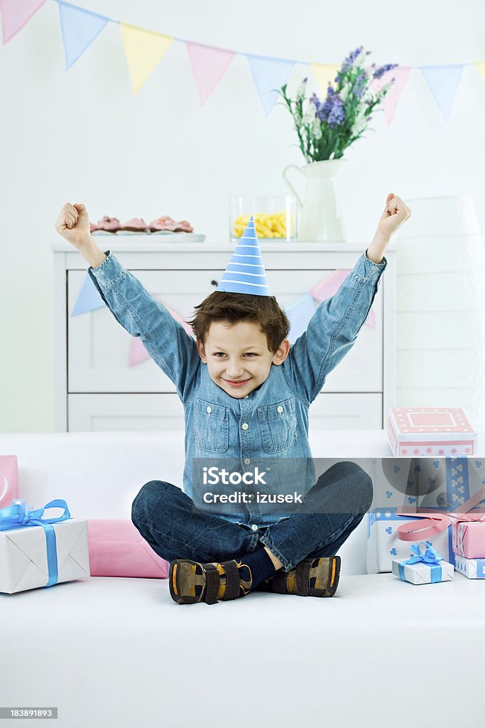 Happy birthday "Cute little boy wearing party hat sitting cross-legged on sofa among birthday presents, raising his hand and smiling." Birthday Stock Photo