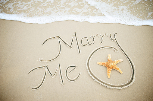 Marry Me message handwritten in smooth brown sand with starfish is bordered by a fresh foamy wave