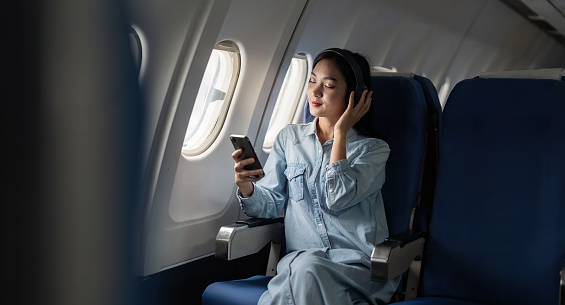 Young female listening song during flight in first class cabin using smartphone, woman entertain on airplane board enjoy music in headphone from smartphone.