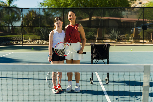 Happy teen girl and hispanic woman coach smiling holding rackets on the tennis court after practice exercises