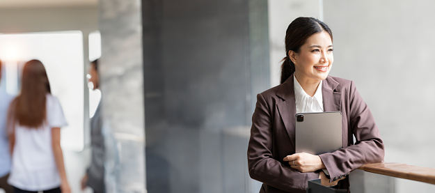 Smiling confident business leader with tablet standing in office, looking up outside.
